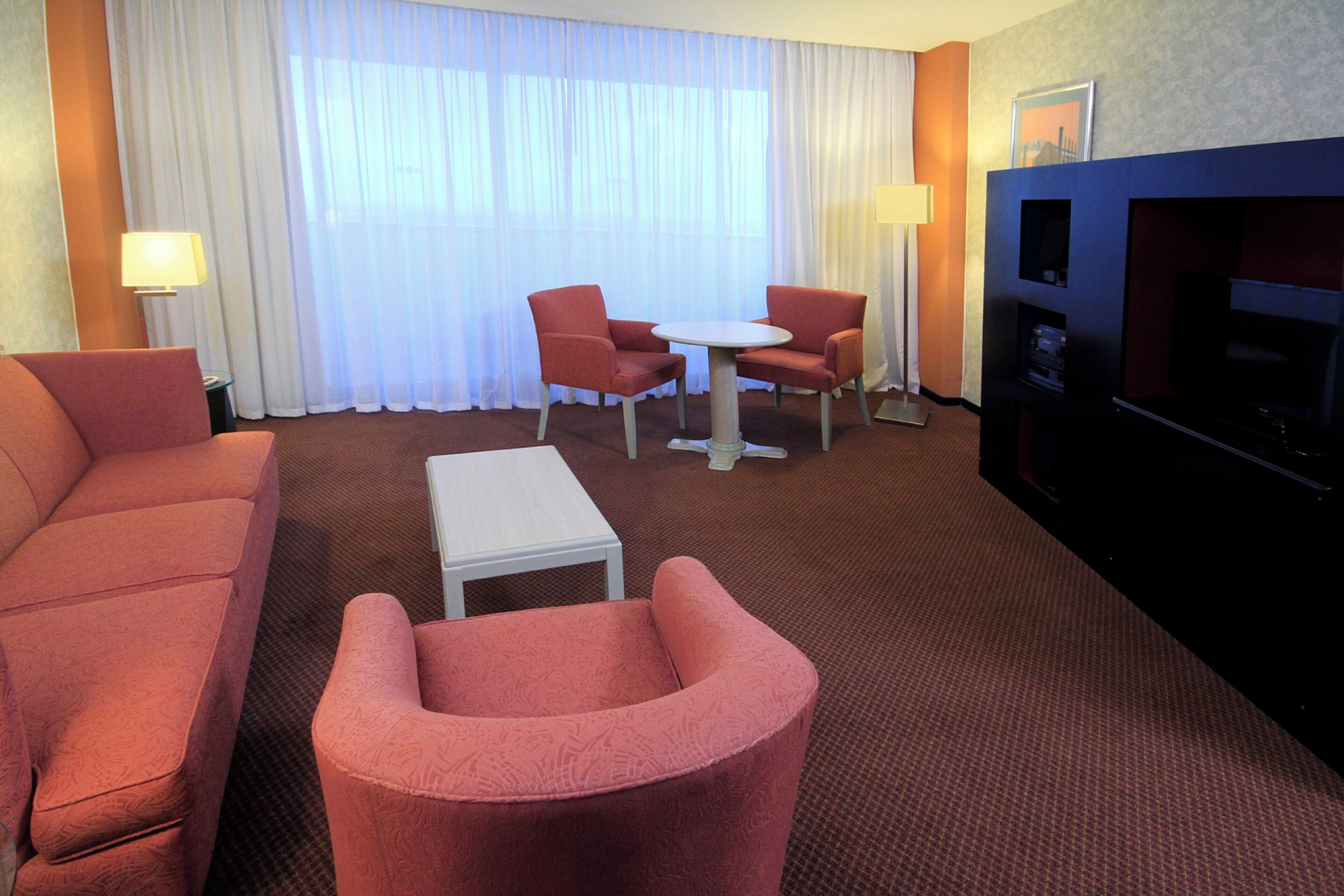 A hotel room with a sofa, two armchairs, a small table, a TV, and a window with curtains.
