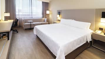 Habitación de hotel con una cama grande, un sofá, un escritorio y una silla. Hay dos lámparas y un televisor en la pared.