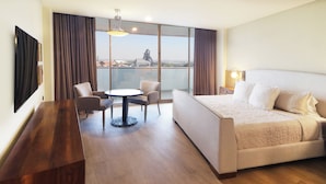 A hotel room with a large bed, a dining table with chairs, a TV, and a view of a cityscape.