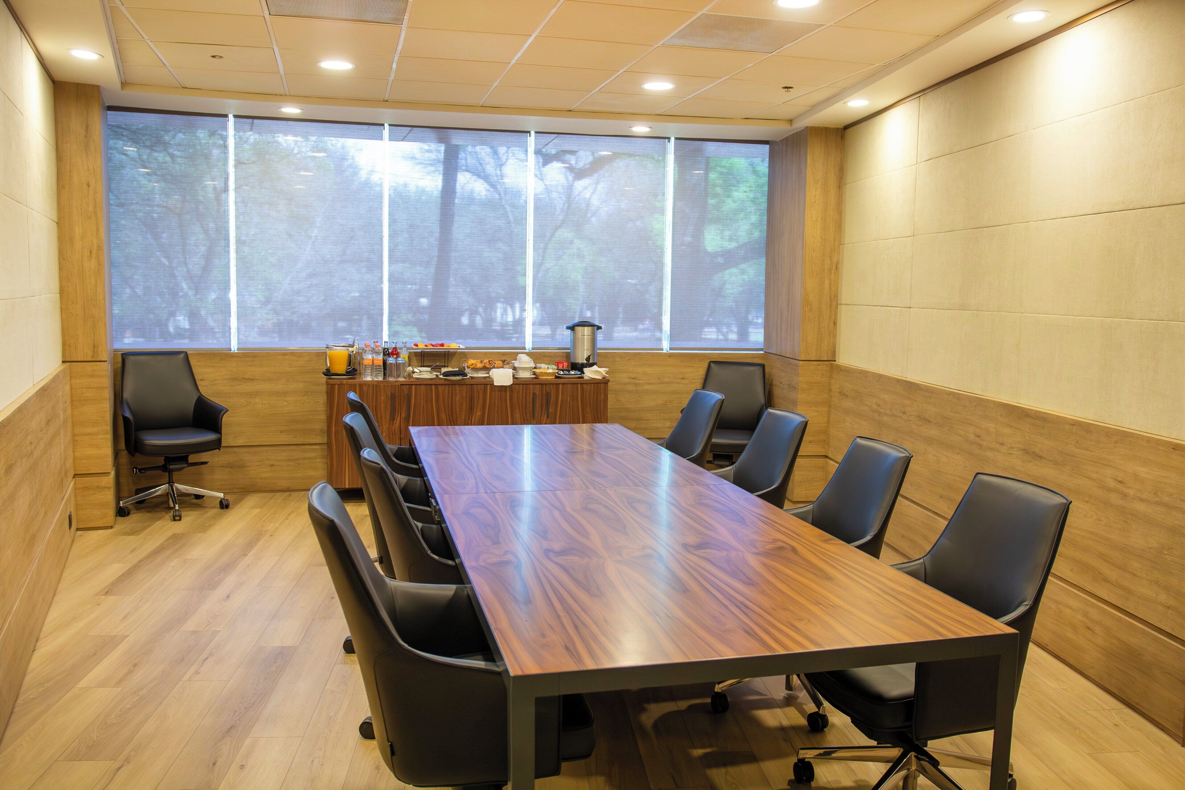 Un salón de conferencias con una mesa larga, seis sillas, una pantalla de proyector y un ventanal con vista al bosque.
