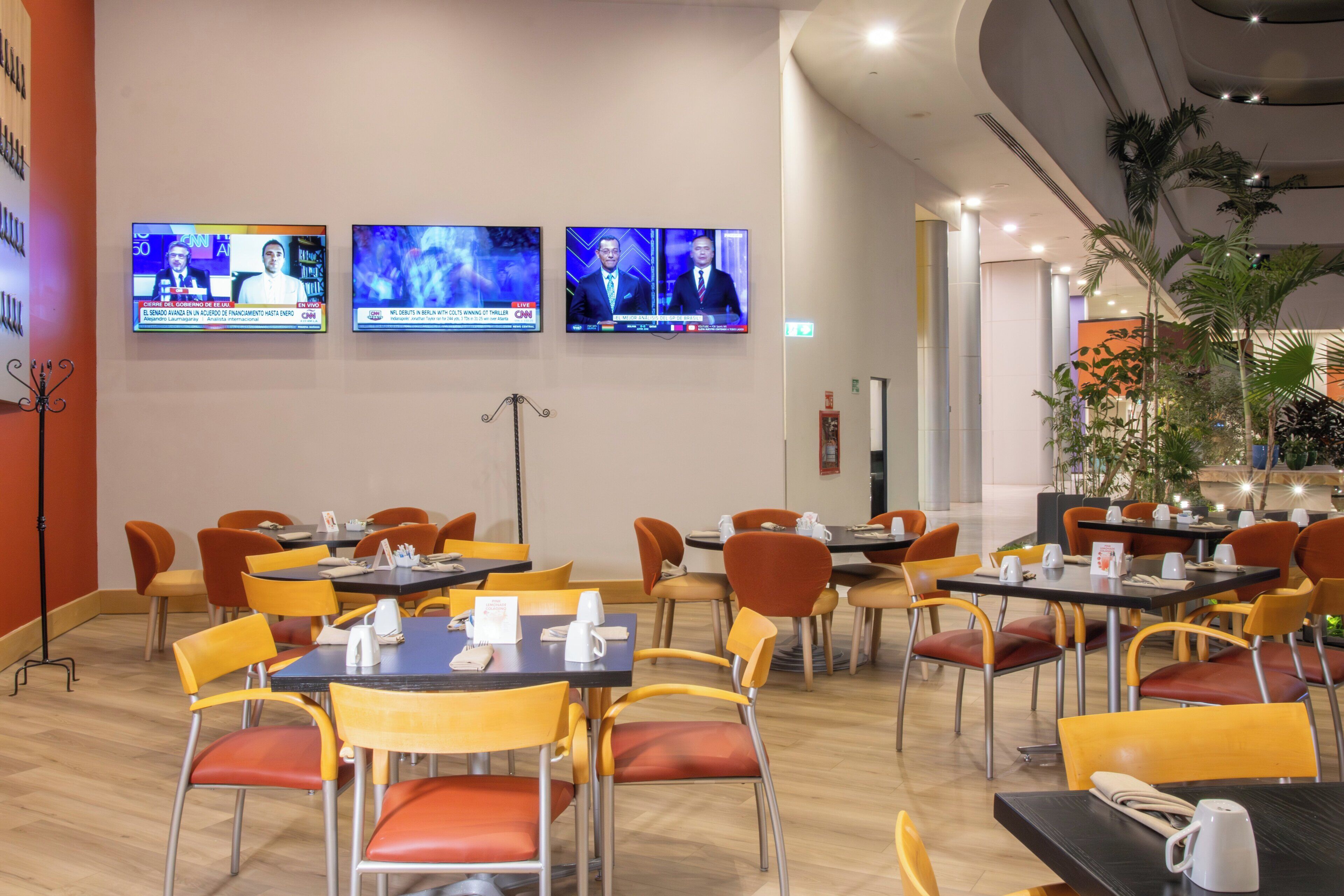 A dining area with multiple tables and chairs, a television mounted on the wall displaying news, and indoor plants.