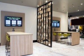 Uma área de recepção moderna com balcão de madeira, assentos de bar e duas telas digitais exibindo códigos QR para serviços.