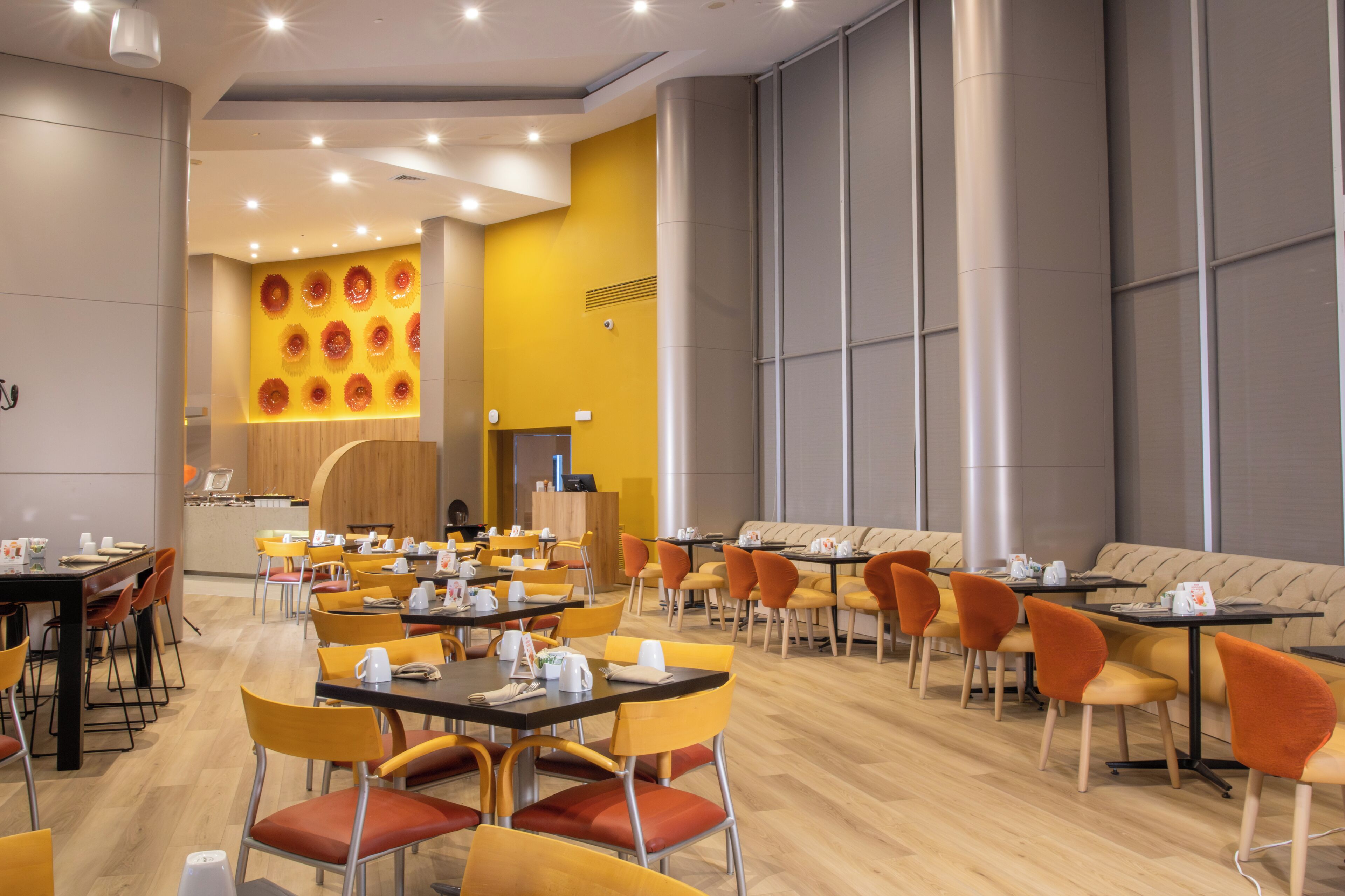 Un espace repas moderne avec des chaises jaunes et orange, des tables en bois et un mur d’accent jaune orné de motifs circulaires.