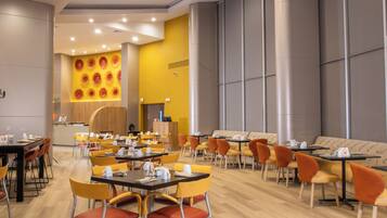 Un espace repas moderne avec des chaises jaunes et orange, des tables en bois et un mur d’accent jaune orné de motifs circulaires.
