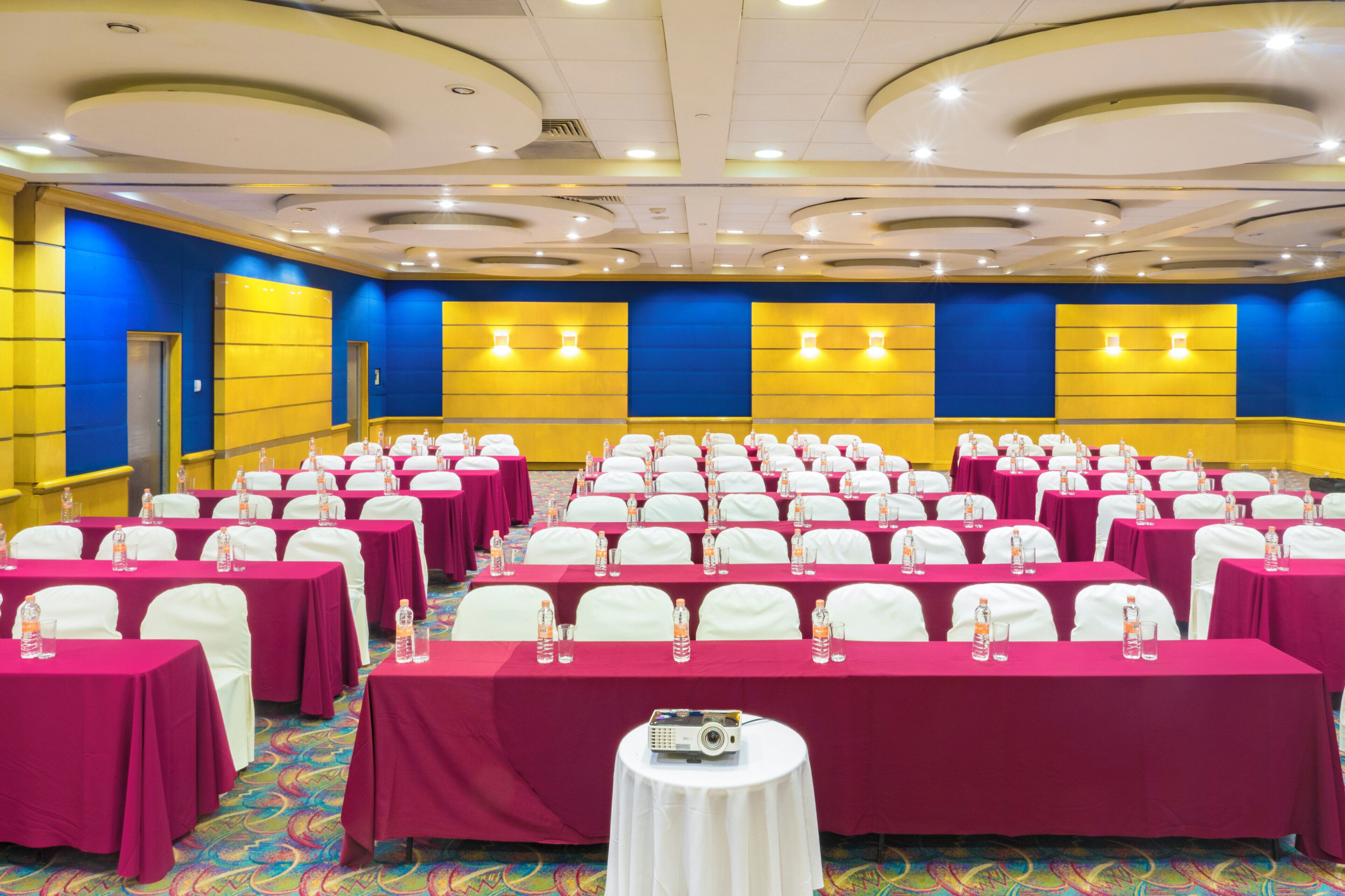 A conference room with rows of tables, each with a white tablecloth and water bottles, in a room with blue and yellow walls and a patterned carpet.