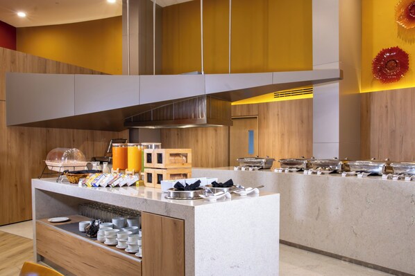 Disposição de buffet com diversos alimentos e recipientes, um espaço para café e uma área de estar com cadeiras.