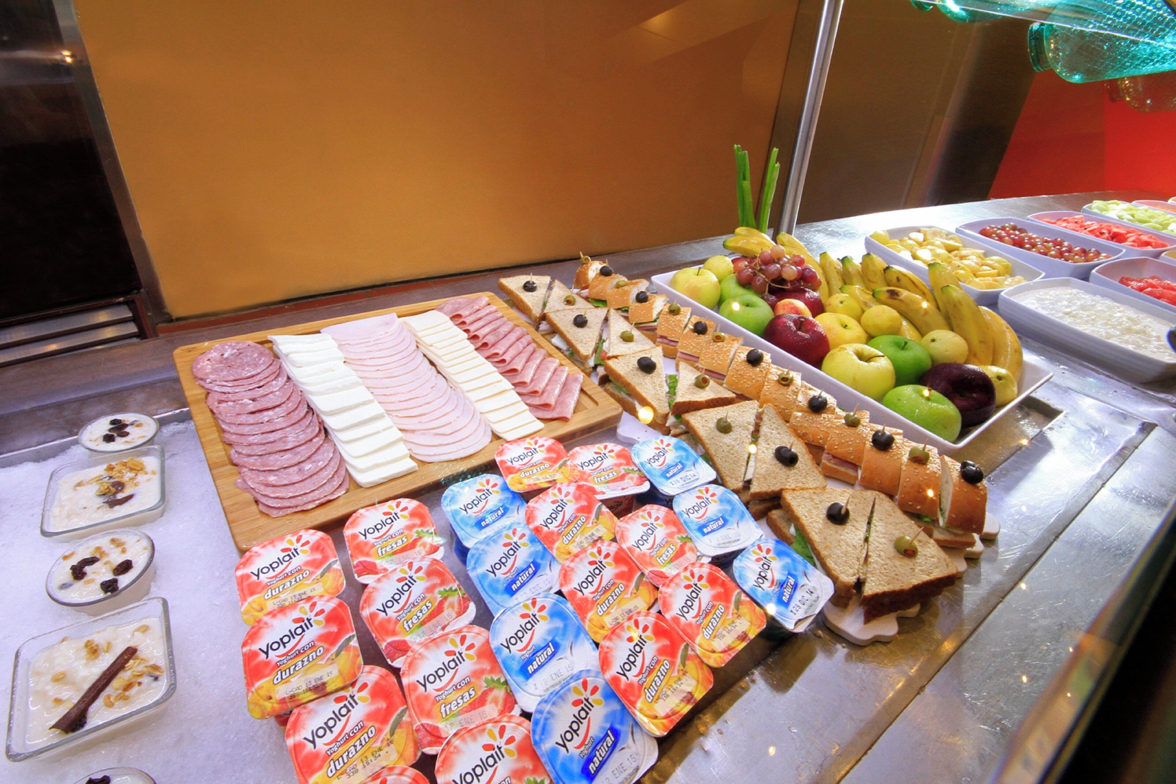 Un bufé con diversas opciones de comida, incluyendo fiambres, quesos, sándwiches, frutas y yogures envasados.