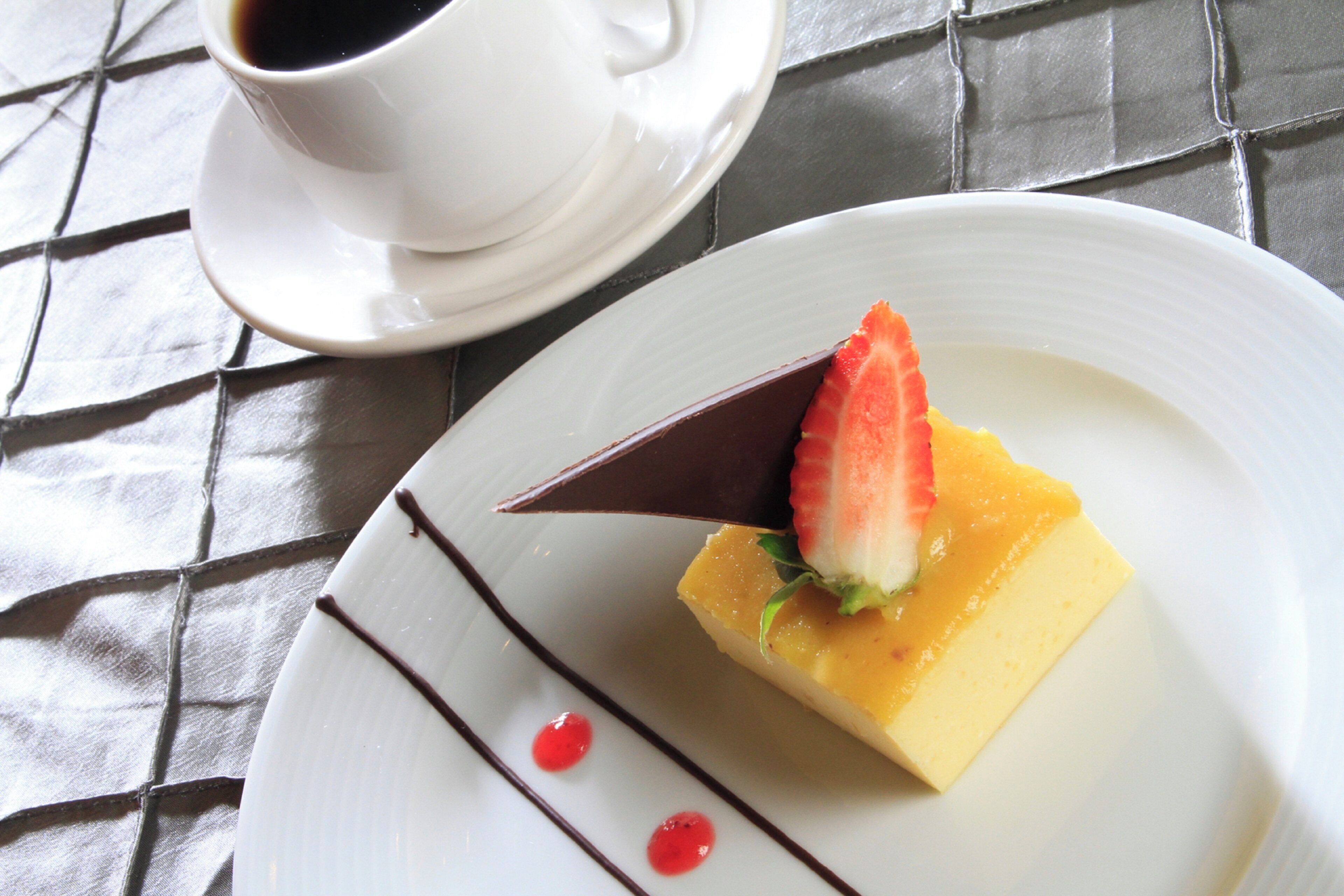 Un postre en un plato blanco con decoración de chocolate, una fresa y un topping amarillo, acompañado de una taza de café sobre un platito.