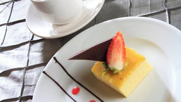 Un postre en un plato blanco con decoración de chocolate, una fresa y un topping amarillo, acompañado de una taza de café sobre un platito.