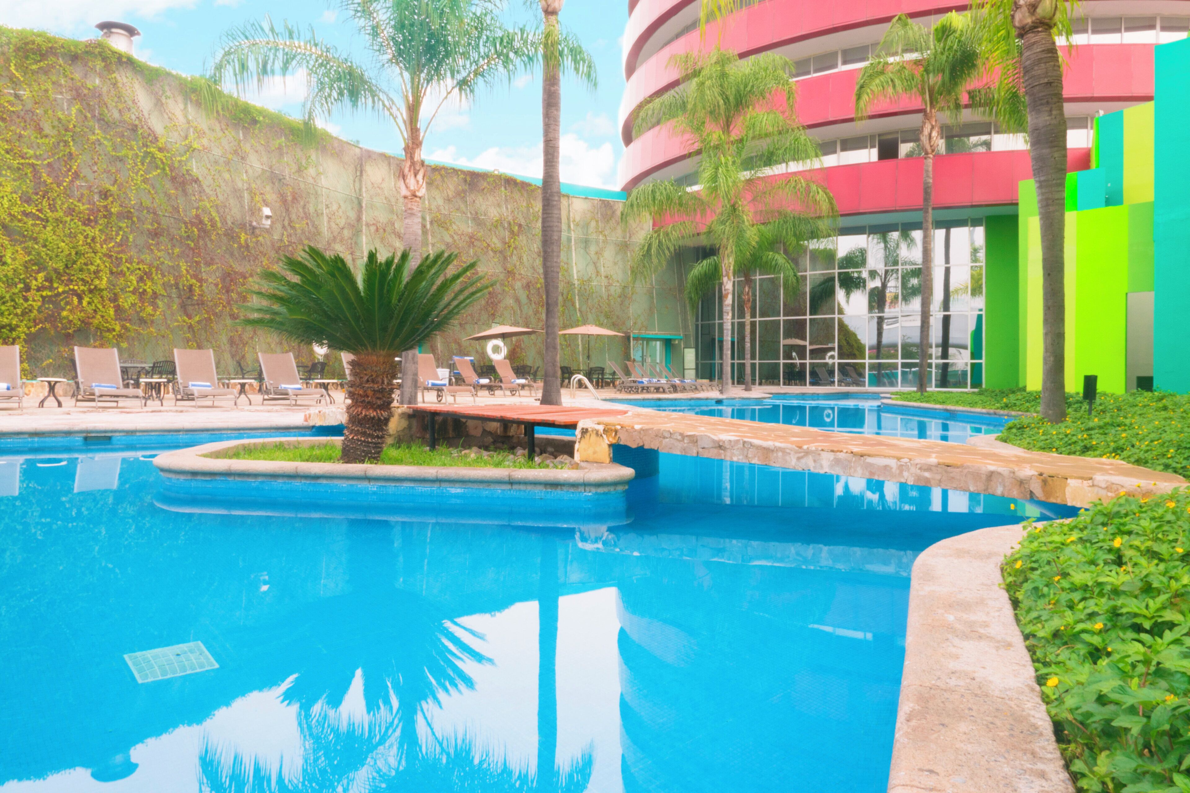 Piscina con sillones de descanso y sombrillas, rodeada de follaje y un edificio con fachada roja y verde.