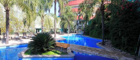 Una piscina con borde de piedra, rodeada de palmeras y un edificio con fachada roja y blanca al fondo.