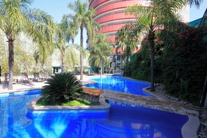 A swimming pool with a stone edge, surrounded by palm trees and a building with a red and white facade in the background. - Holiday Inn Monterrey - Parque Fundidora by IHG (Monterrey)