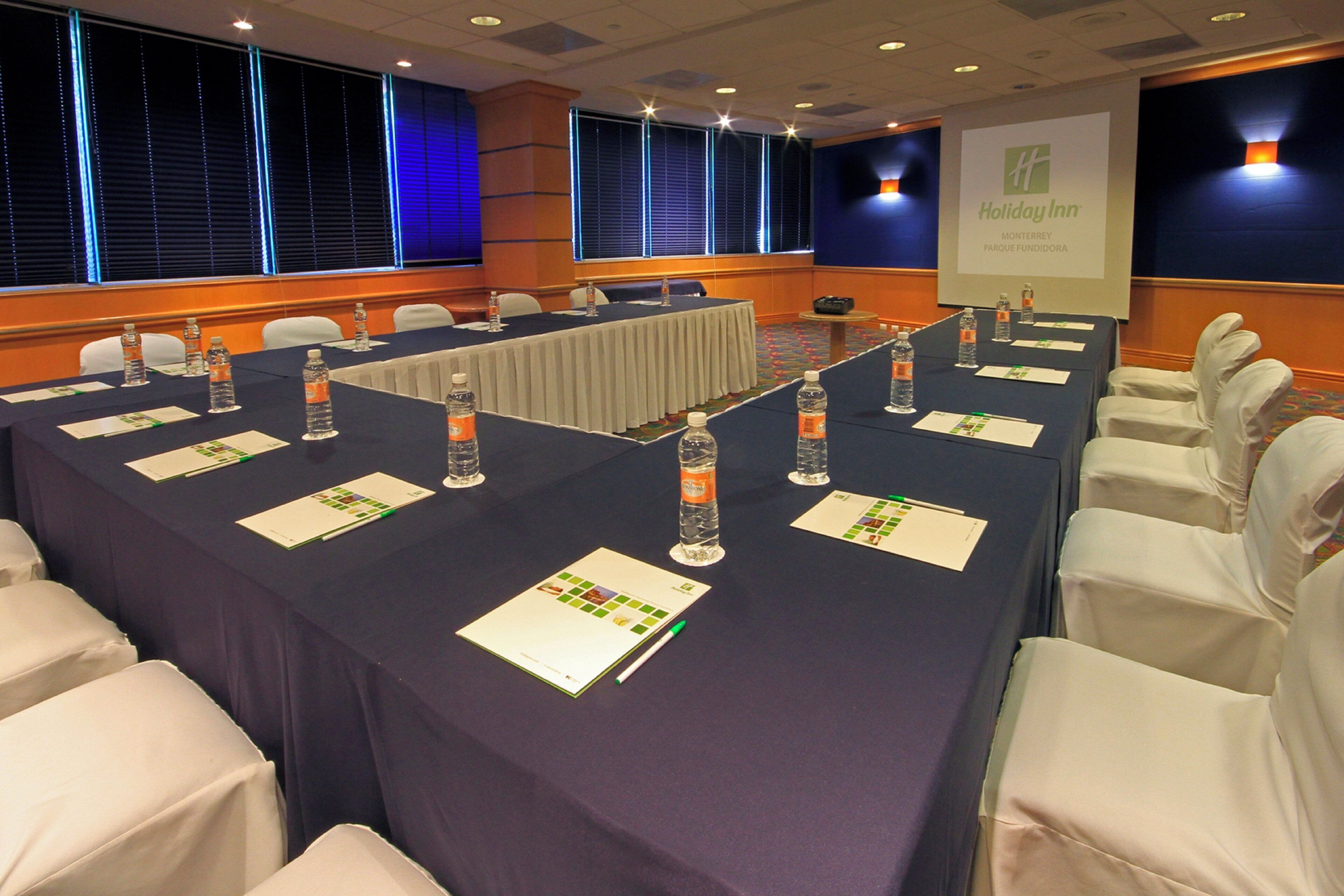 Un salón de conferencias con una larga mesa, sillas, botellas de agua, tarjetas de identificación y una pantalla para presentaciones.