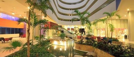 A spacious indoor atrium with a central water feature, surrounded by greenery and seating areas.