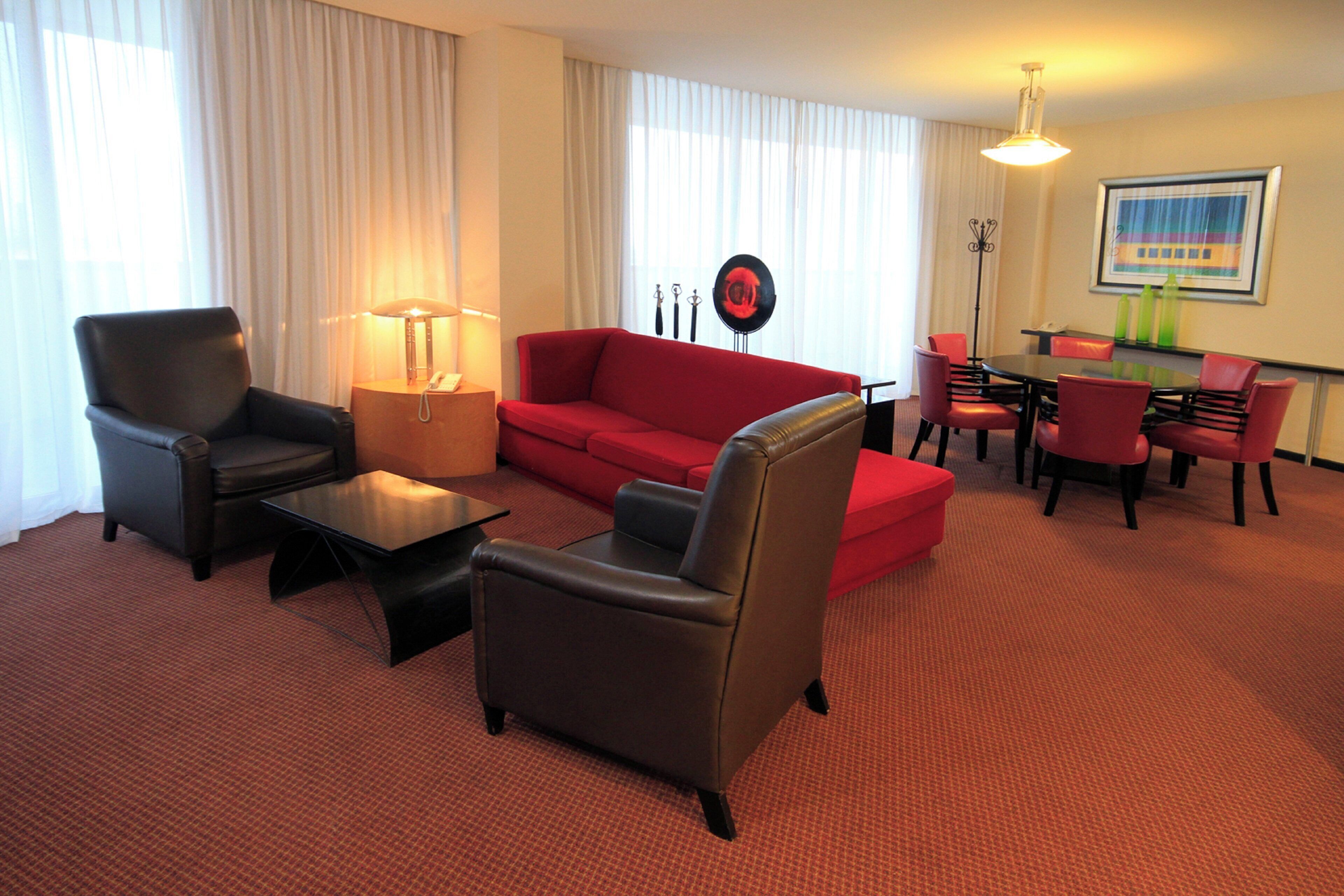 A hotel room with a red sofa, two armchairs, a coffee table, a dining table with chairs, a lamp, and a painting on the wall.