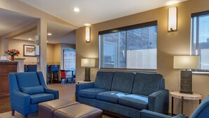 Un salon moderne avec un canapé bleu, un fauteuil assorti et une table basse. On y trouve deux tables d’appoint avec des lampes, une cheminée et une grande fenêtre qui laisse entrer la lumière naturelle.