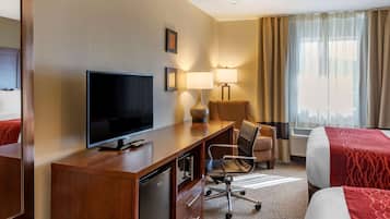 Een hotelkamer met een flatscreen televisie, een bureau met een lamp, een stoel, een bed met een rode deken, en een raam met gordijnen.
