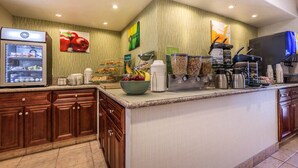 A hotel breakfast area with a refrigerator, coffee machines, and a bowl of fruit.