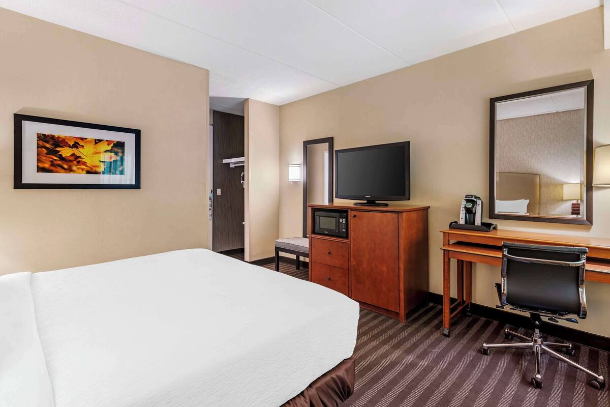 A hotel room with a bed, a desk with a computer, a television, a mirror, and a framed picture on the wall.