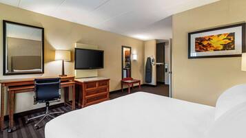 A hotel room with a large bed, a desk with a chair, a flat-screen TV, a mirror, a small table, and a framed picture on the wall.