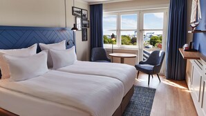 Habitación de hotel con dos camas, una mesita redonda pequeña, una silla y vistas a la ciudad a través de amplias ventanas.