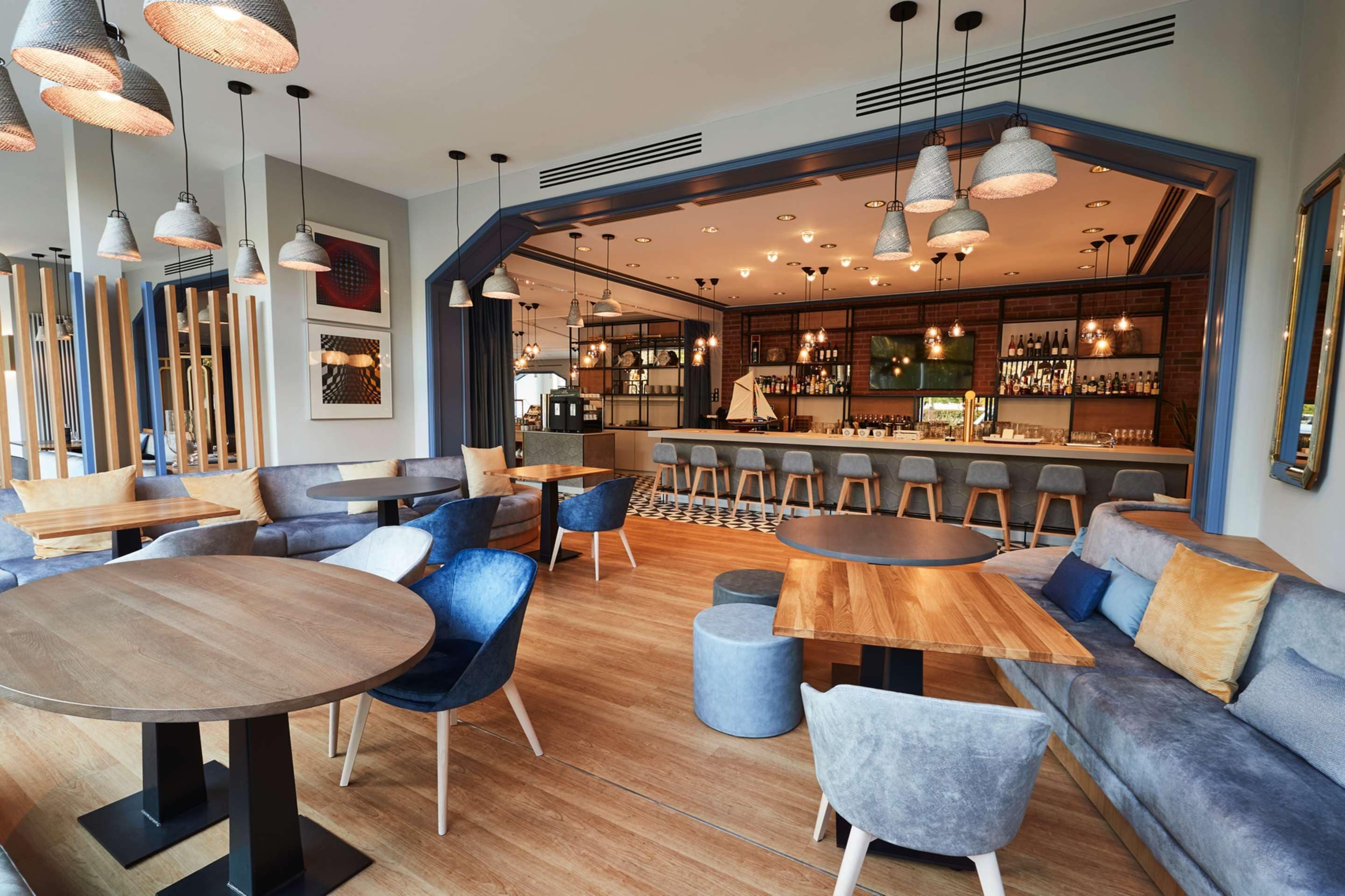 A modern bar area with wooden tables, blue chairs, and a brick wall backdrop.