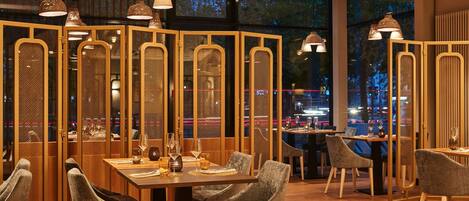 Un restaurante con muebles de madera, iluminación colgante y vistas de la ciudad iluminada por la noche.