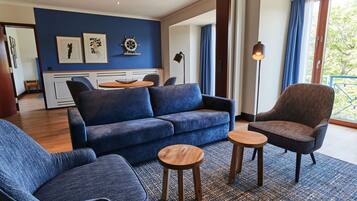 A modern living room with a blue sofa, two armchairs, and a coffee table. There are framed artworks on the wall, a floor lamp, and a view of the outdoors through a large window.