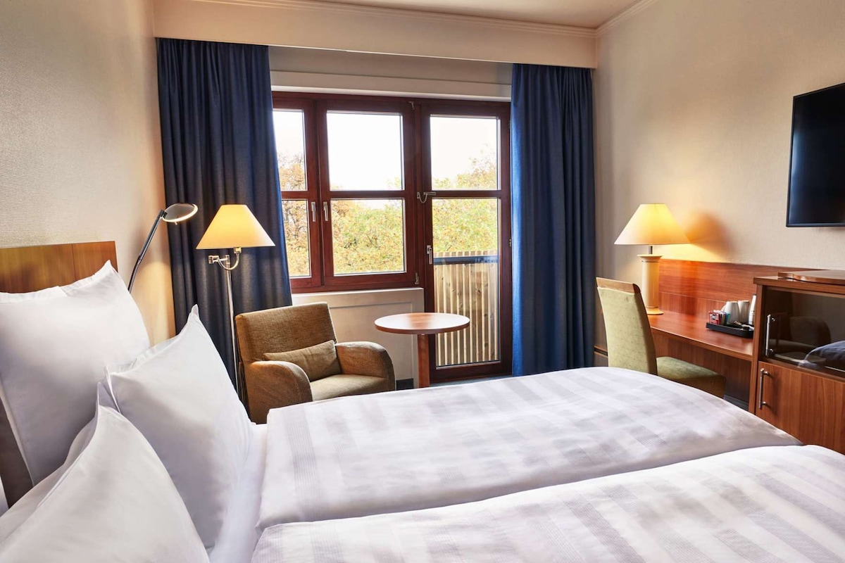 A hotel room with a bed, two chairs, a small table, a desk with a lamp, and a TV.
