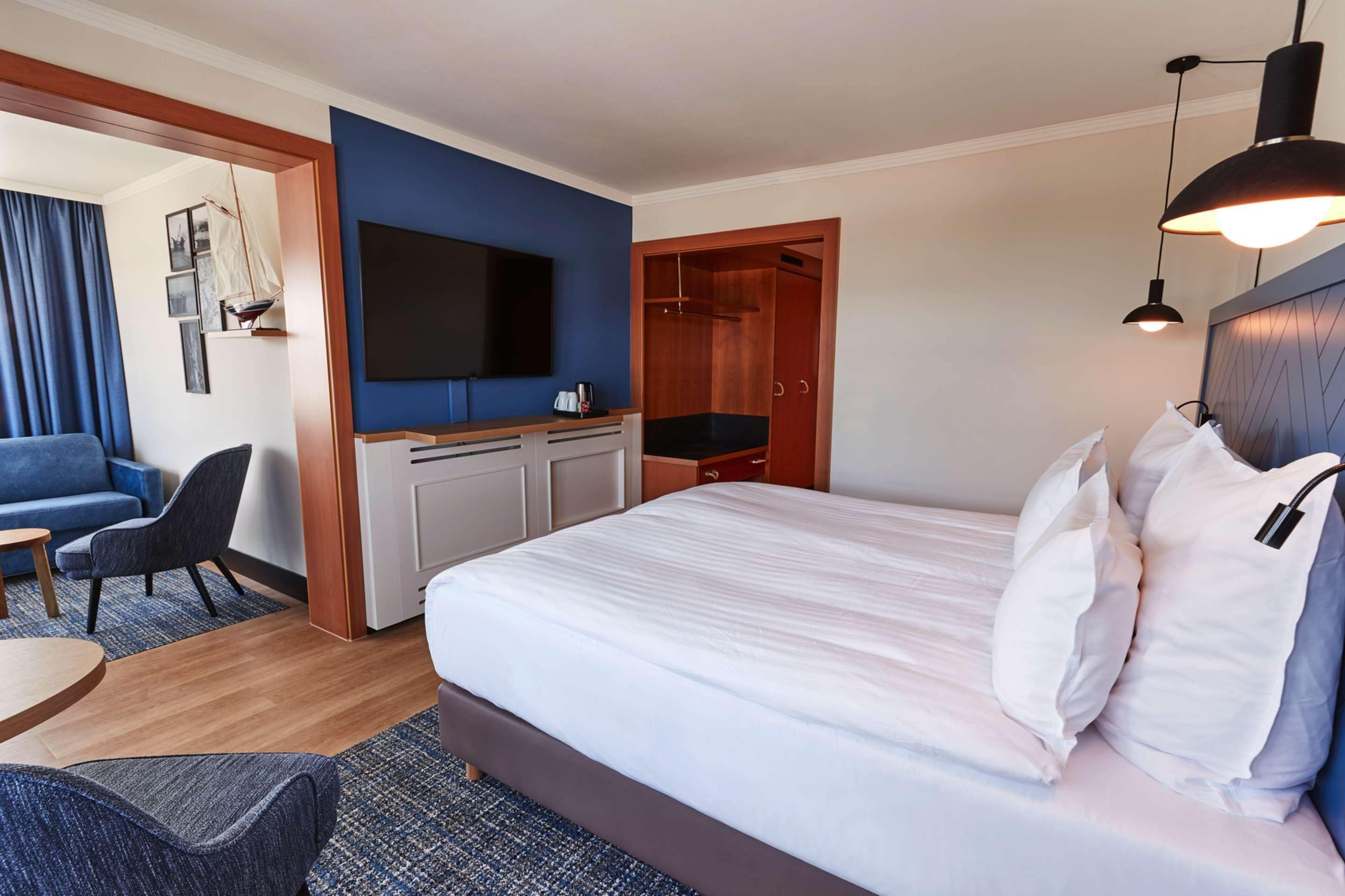 Ein Hotelzimmer mit Bett, Fernseher, Schreibtisch, Sessel, Kleiderschrank und einem Fenster mit Vorhängen.