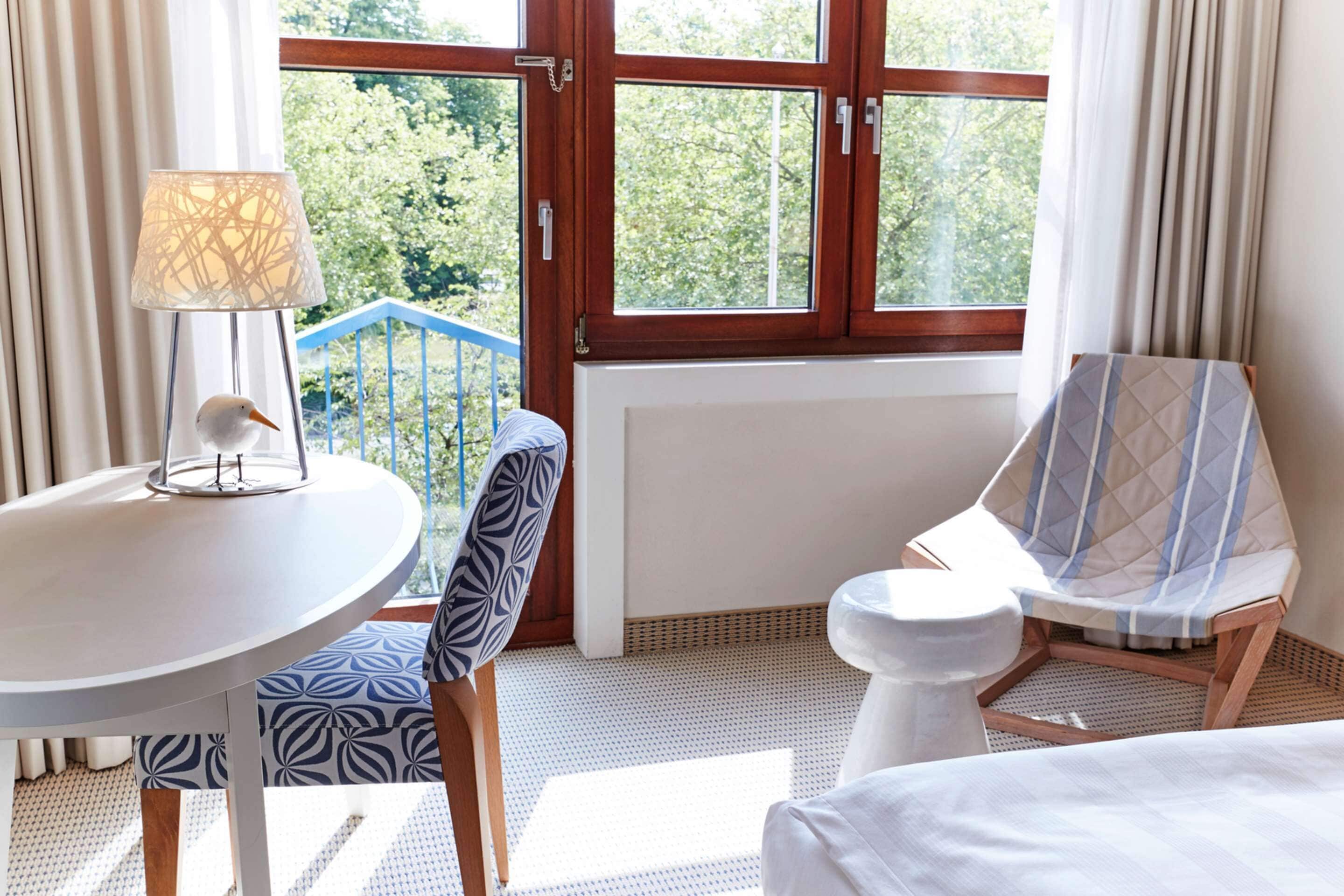 Ein Hotelzimmer mit großem Fenster, einem runden Tisch mit Lampe, einem gemusterten Sessel, einer weißen Toilette und einem Bett.