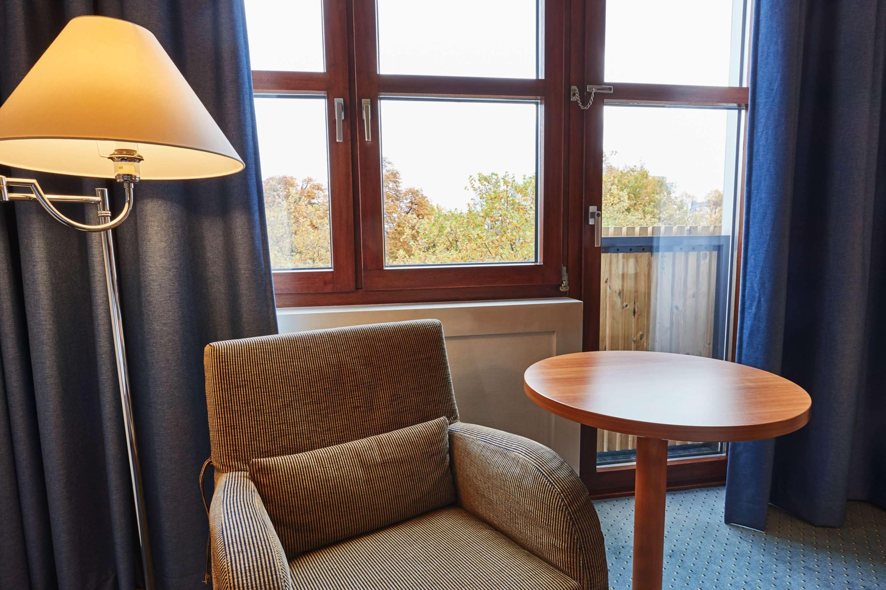 Een kamer met een bank, een ronde tafel, een lamp en een raam met uitzicht op bomen en een houten hek.