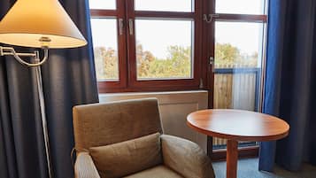 Een kamer met een bank, een ronde tafel, een lamp en een raam met uitzicht op bomen en een houten hek.