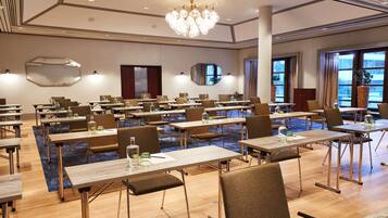 A conference room with multiple tables and chairs, a large chandelier, and large windows with curtains.