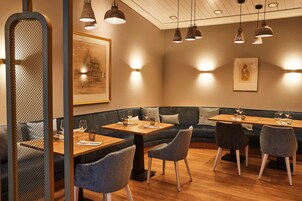 A restaurant interior with wooden tables, cushioned seating, pendant lighting, and artwork on the walls.