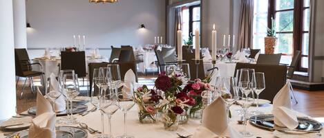 Un salón de banquetes con mesas redondas dispuestas para un evento formal, con manteles blancos, cubertería y centros de mesa con velas.