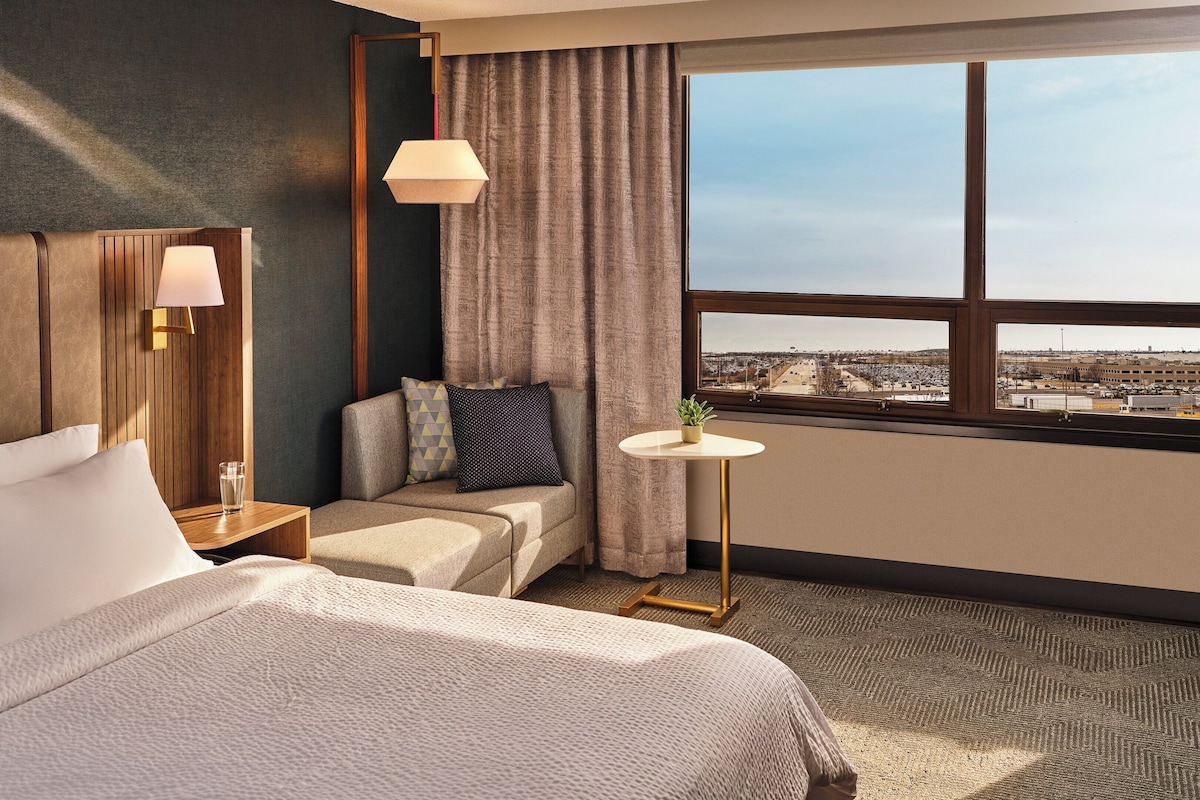 A hotel room with a bed, a seating area, a small table with a plant, a lamp, and a view of the city through a large window.