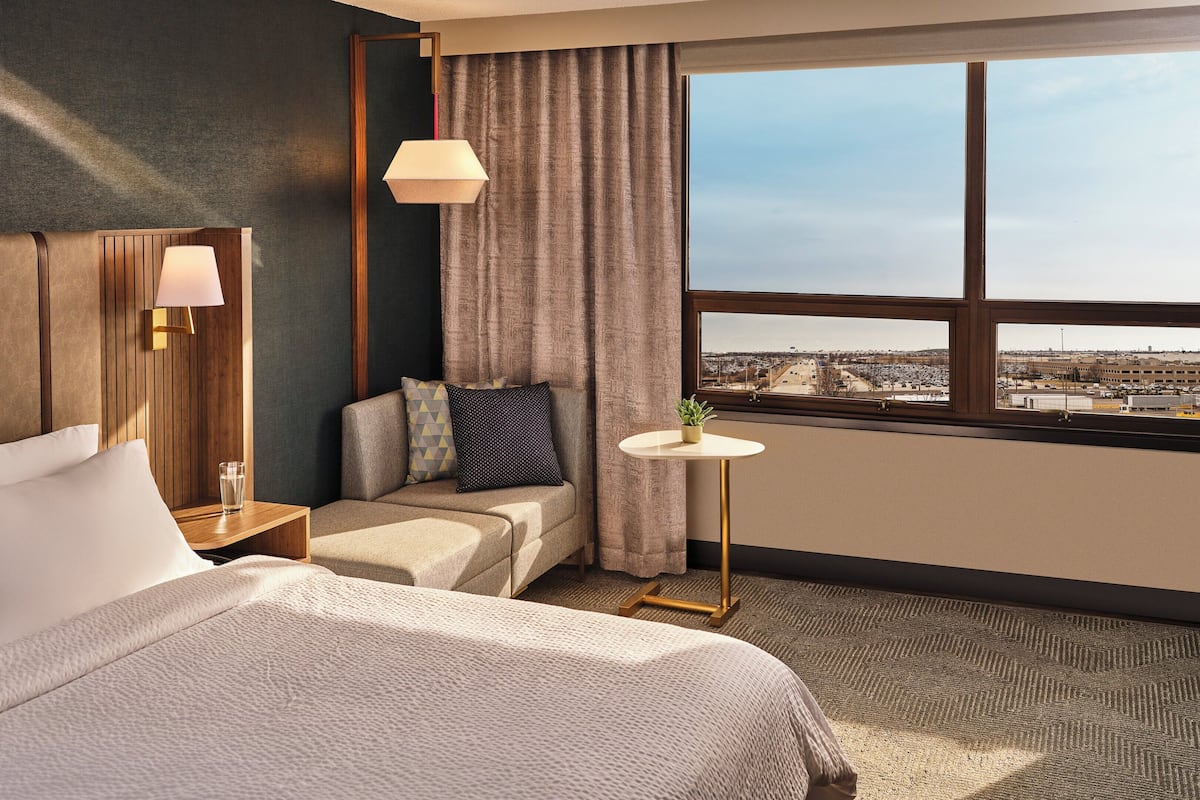 Ein Hotelzimmer mit Bett, einer Sitzecke, einem kleinen Tisch mit Pflanze, einer Lampe und Blick auf die Stadt durch ein großes Fenster.
