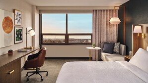 A hotel room with a large bed, a desk, a chair, a small table with a plant, and a view of the city through the window. - Holiday Inn Cleveland Airport West by IHG (North Olmsted)
