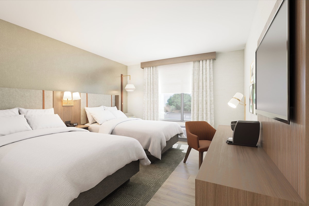 Ein Hotelzimmer mit zwei Betten, einem Schreibtisch, einem Stuhl, einem Fernseher und einem Fenster mit Vorhängen.