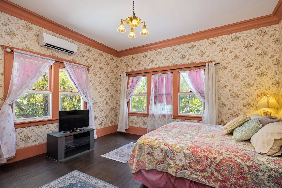 Une chambre à coucher avec un papier peint à motifs floraux, un lit recouvert d’une couvre-lit à motifs, un téléviseur posé sur un meuble et une fenêtre avec des rideaux.