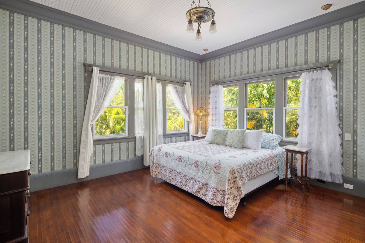 Une chambre à coucher avec un grand lit, deux fenêtres avec des rideaux, un plancher en bois et un lustre.
