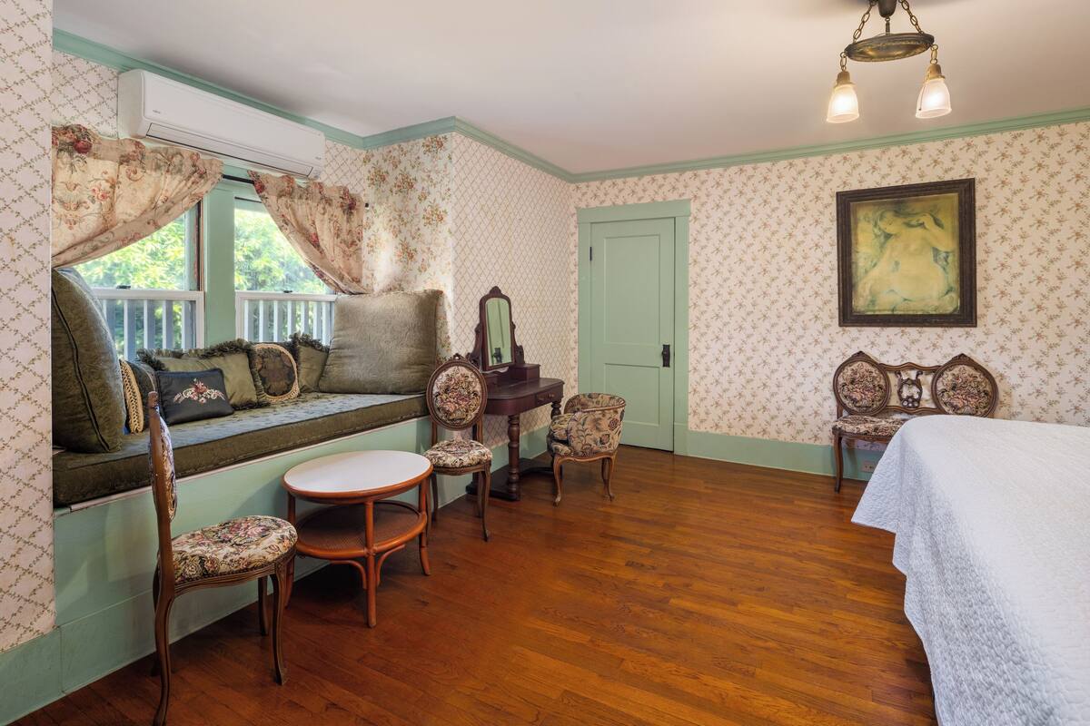 Une pièce avec un plancher en bois, un banc avec des coussins, deux chaises, une table, une porte et un tableau accroché au mur.