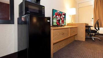 A compact hotel room with a black refrigerator, a wooden desk with a chair, a flat-screen TV, and a lamp.