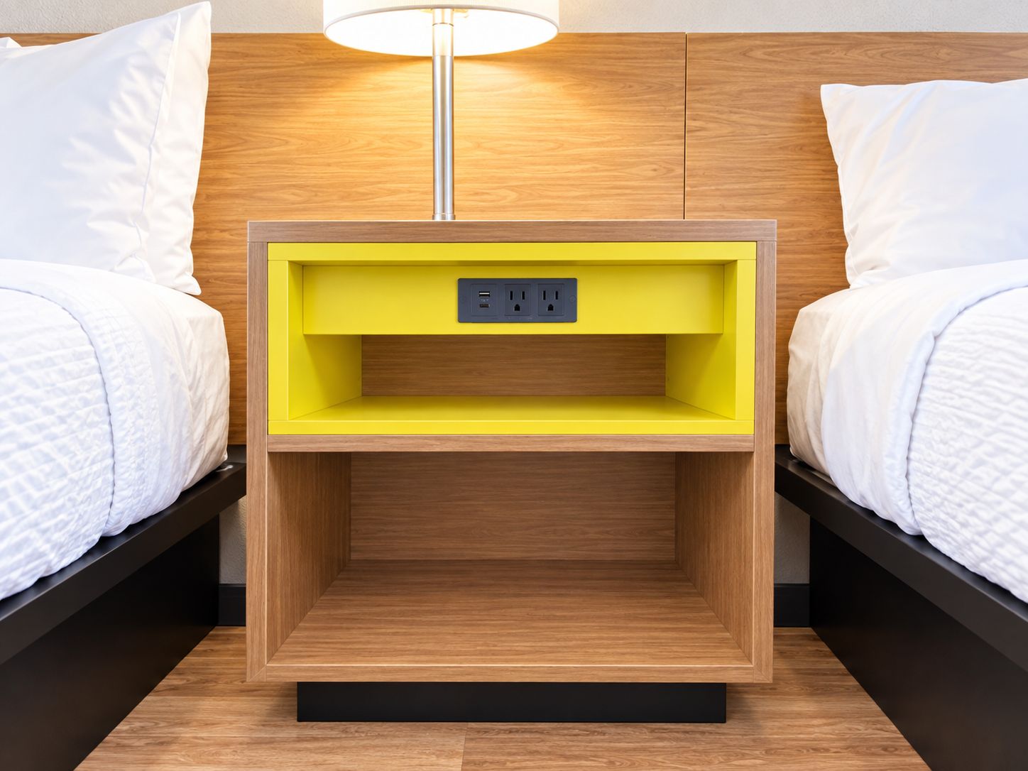 A hotel room with a wooden headboard, a bedside table with a yellow shelf, and a lamp.