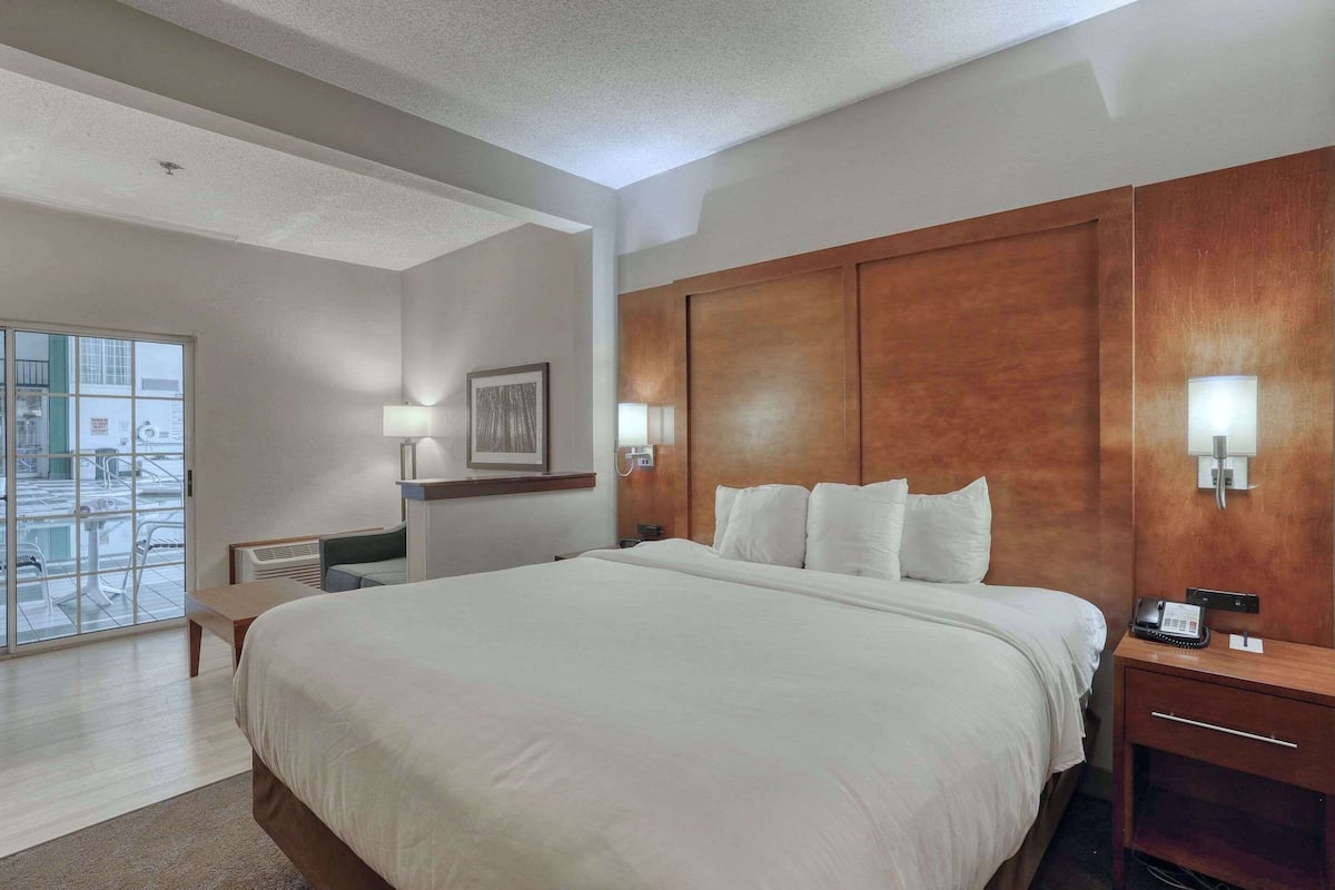 A hotel room with a large bed, a wooden headboard, bedside lamps, a desk, a chair, and a large window with a view of the outside.