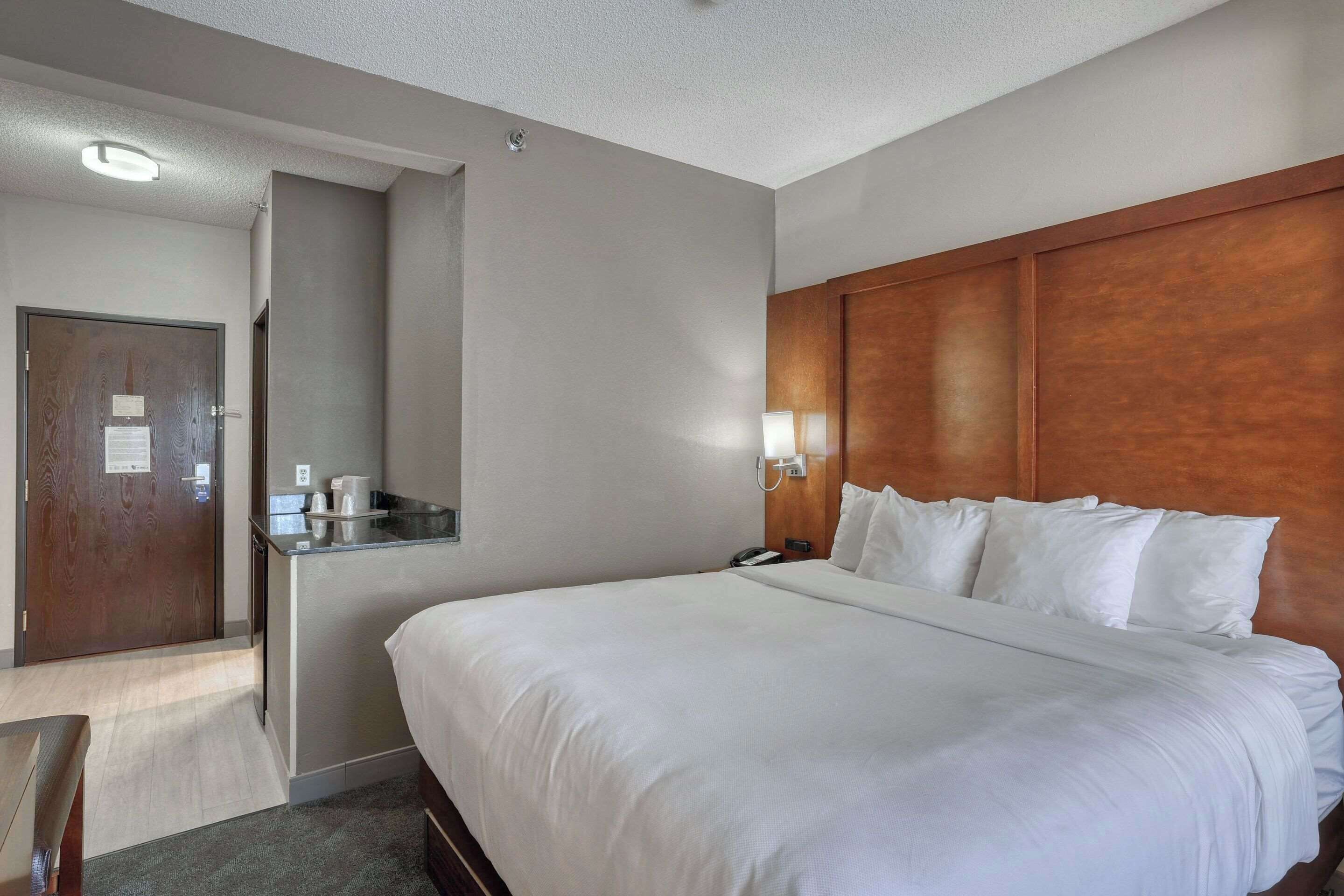A hotel room with a large bed, a wooden headboard, a bedside table with a lamp, and a small desk with a telephone.