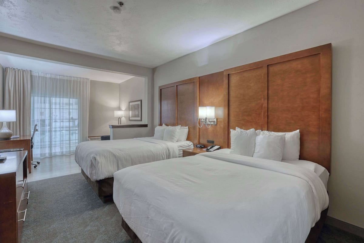 A hotel room with two beds, a desk, and a chair. There is a window with curtains, a lamp, and a framed picture on the wall.