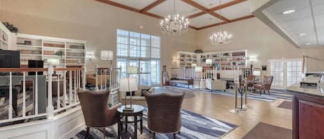 A spacious lobby with high ceilings, wooden beams, and large windows allowing natural light. There are multiple seating areas with chairs and tables, and a grand chandelier hanging from the ceiling.