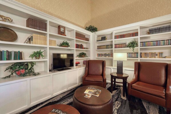 A library with bookshelves, leather armchairs, a round ottoman, and a television.